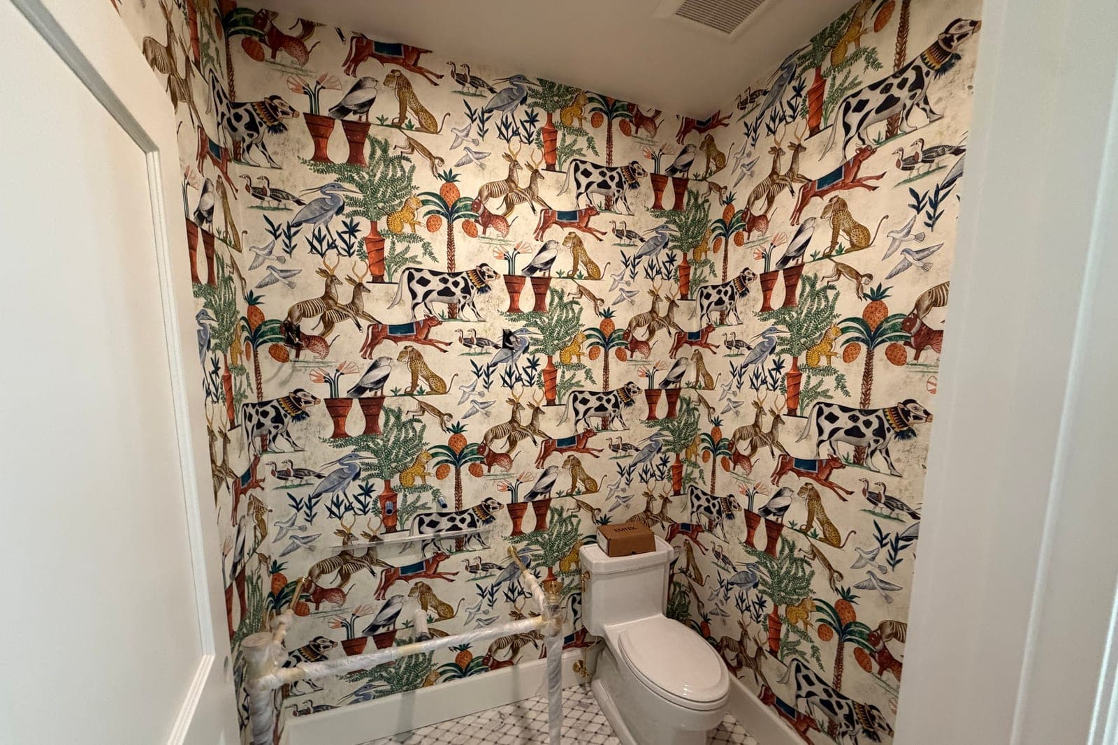 Powder room papered floor to ceiling in a cream Egyptian animal print of cows, herons, cheetahs and palm trees, with a marble mosaic floor