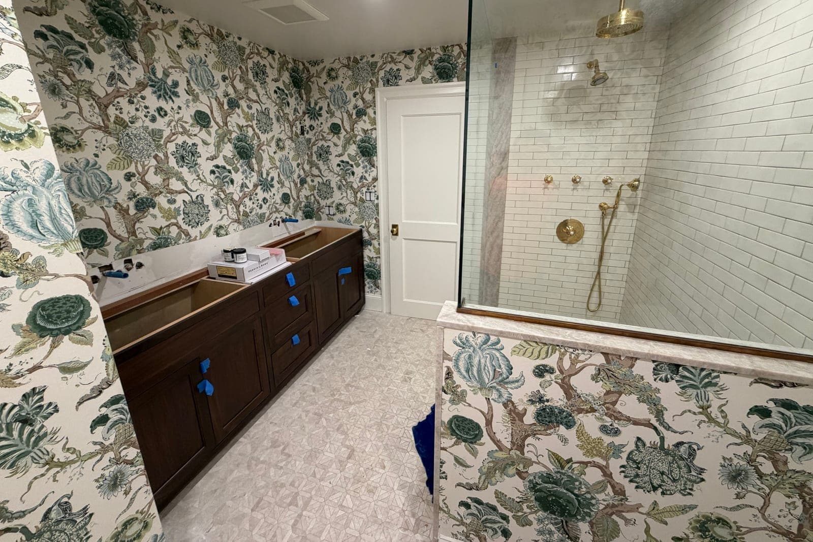 Bathroom in a large blue-green floral chintz on white, with a walnut vanity, a glass shower and brass fittings