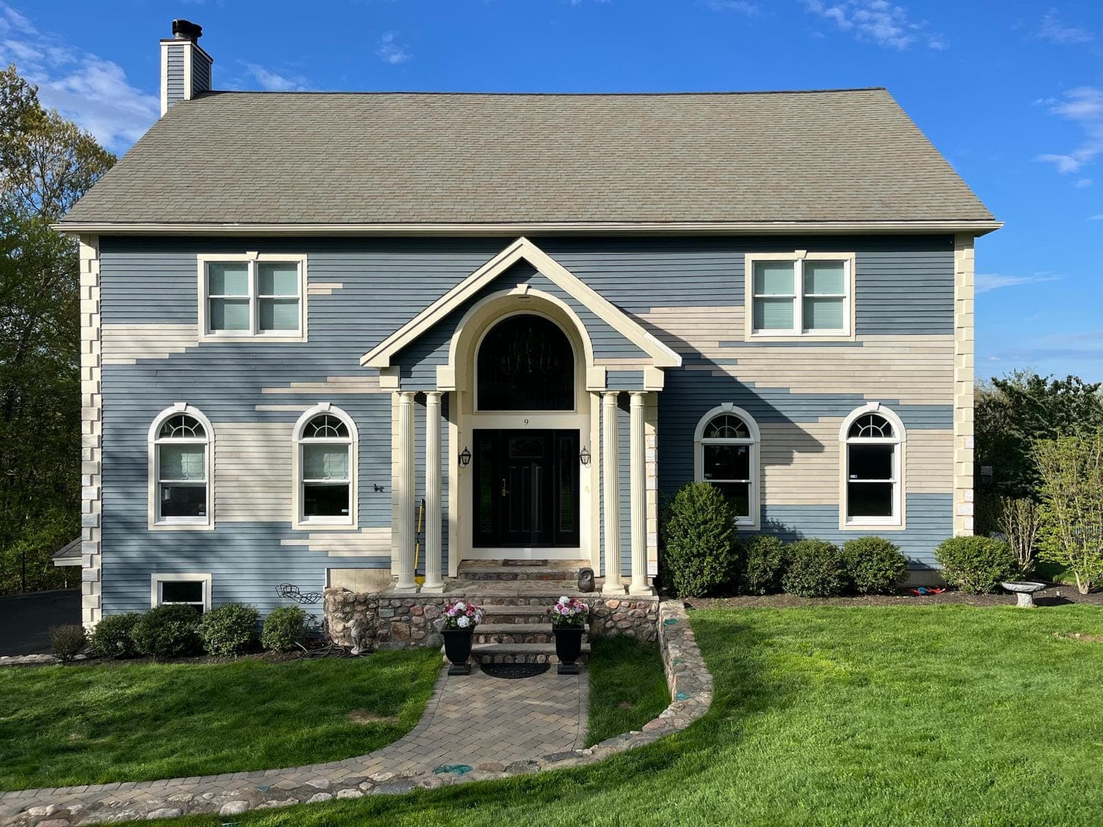 Blue-grey clapboard colonial with a columned portico, new primed clapboard runs showing pale against the old, on a spring afternoon